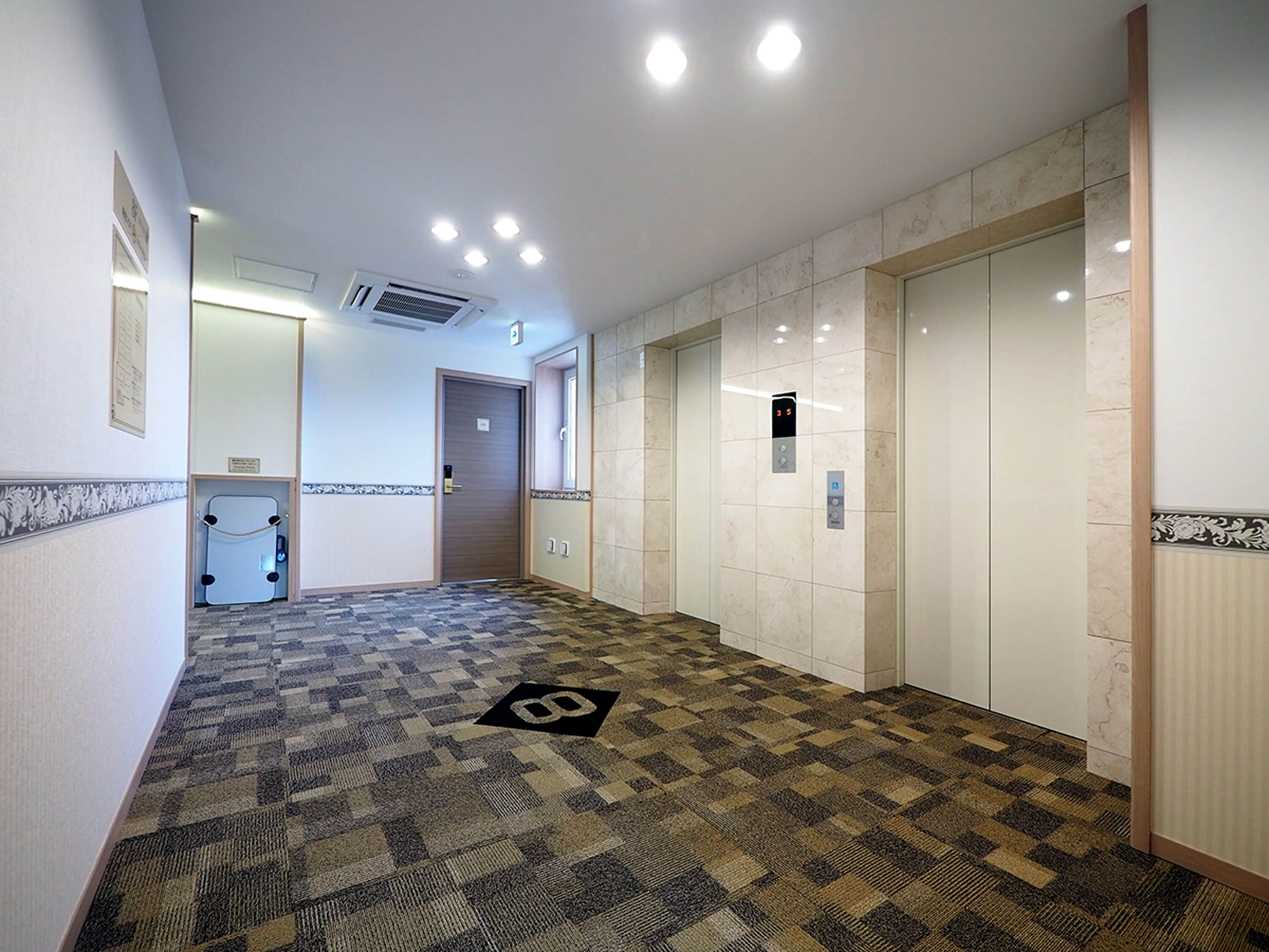 Un pasillo de hotel con alfombra estampada, una puerta con pantalla digital y un ascensor.