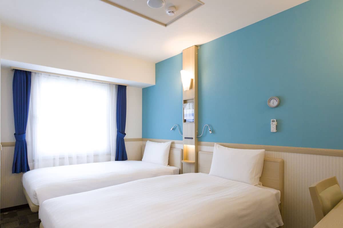 Une chambre d’hôtel avec deux lits, une grande fenêtre avec des rideaux, un radiateur fixé au mur et une horloge.