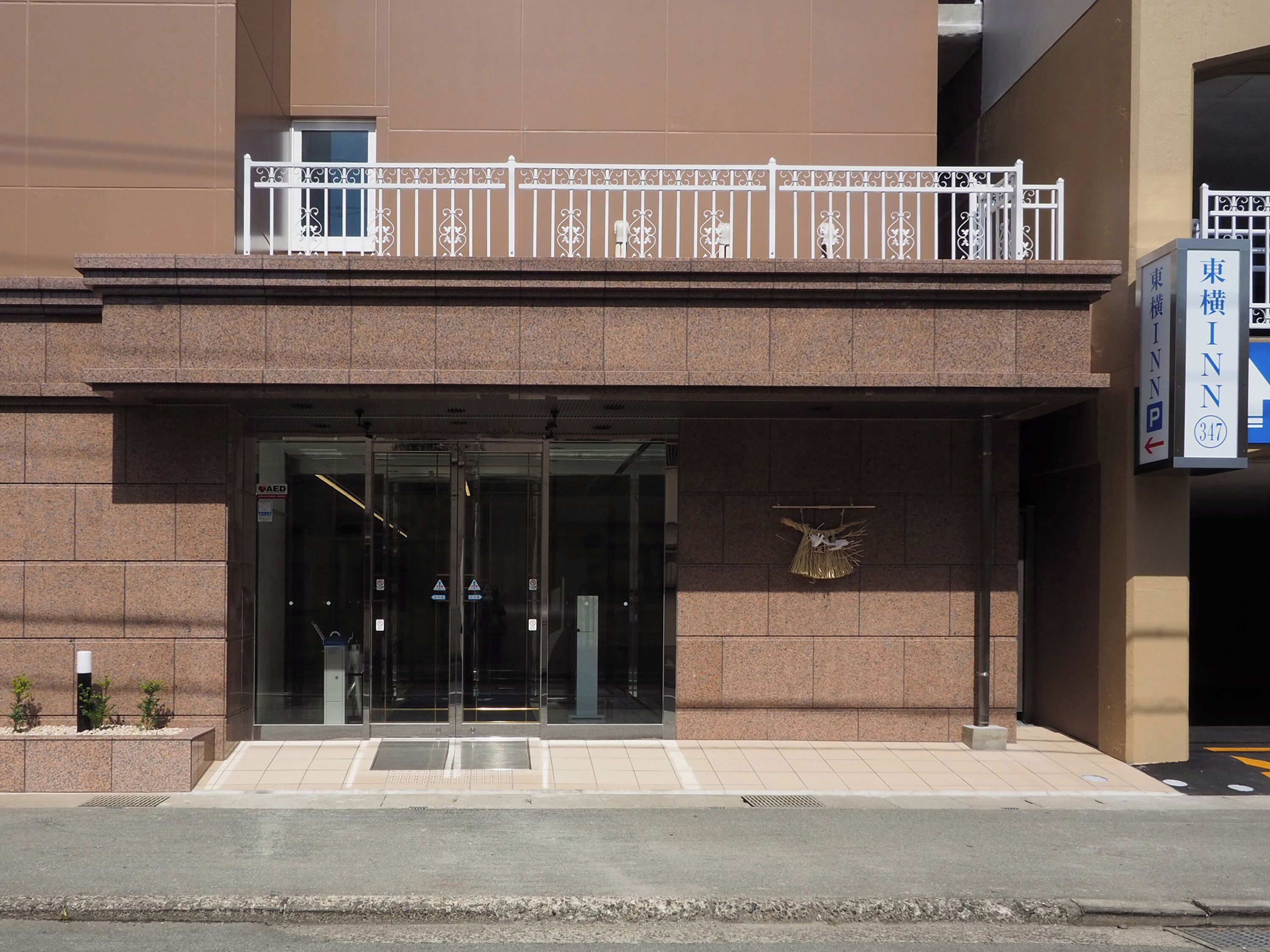 A modern building with a glass entrance, a small outdoor seating area, and a sign indicating the name and address.