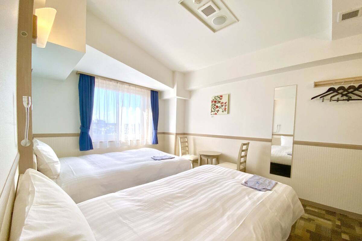 Une chambre d’hôtel avec deux lits, une petite table, une chaise, un miroir et une fenêtre avec des rideaux.