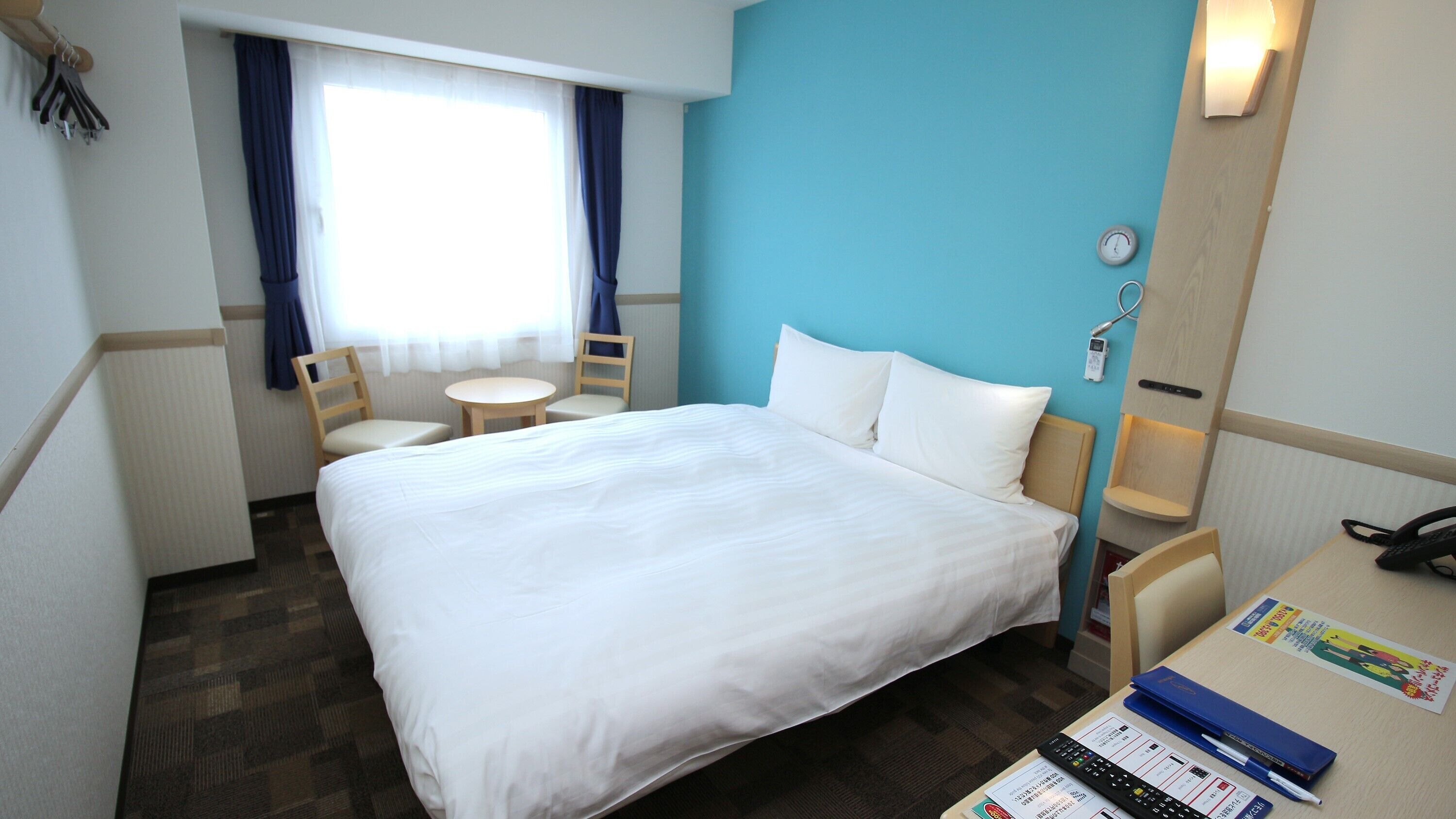 A hotel room with a bed, a desk with a phone and a remote control, a chair, a small table, and a window with curtains.