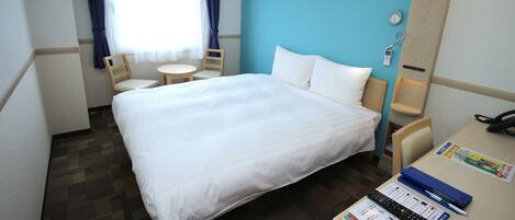 A hotel room with a bed, a desk with a phone and a remote control, a chair, a small table, and a window with curtains.