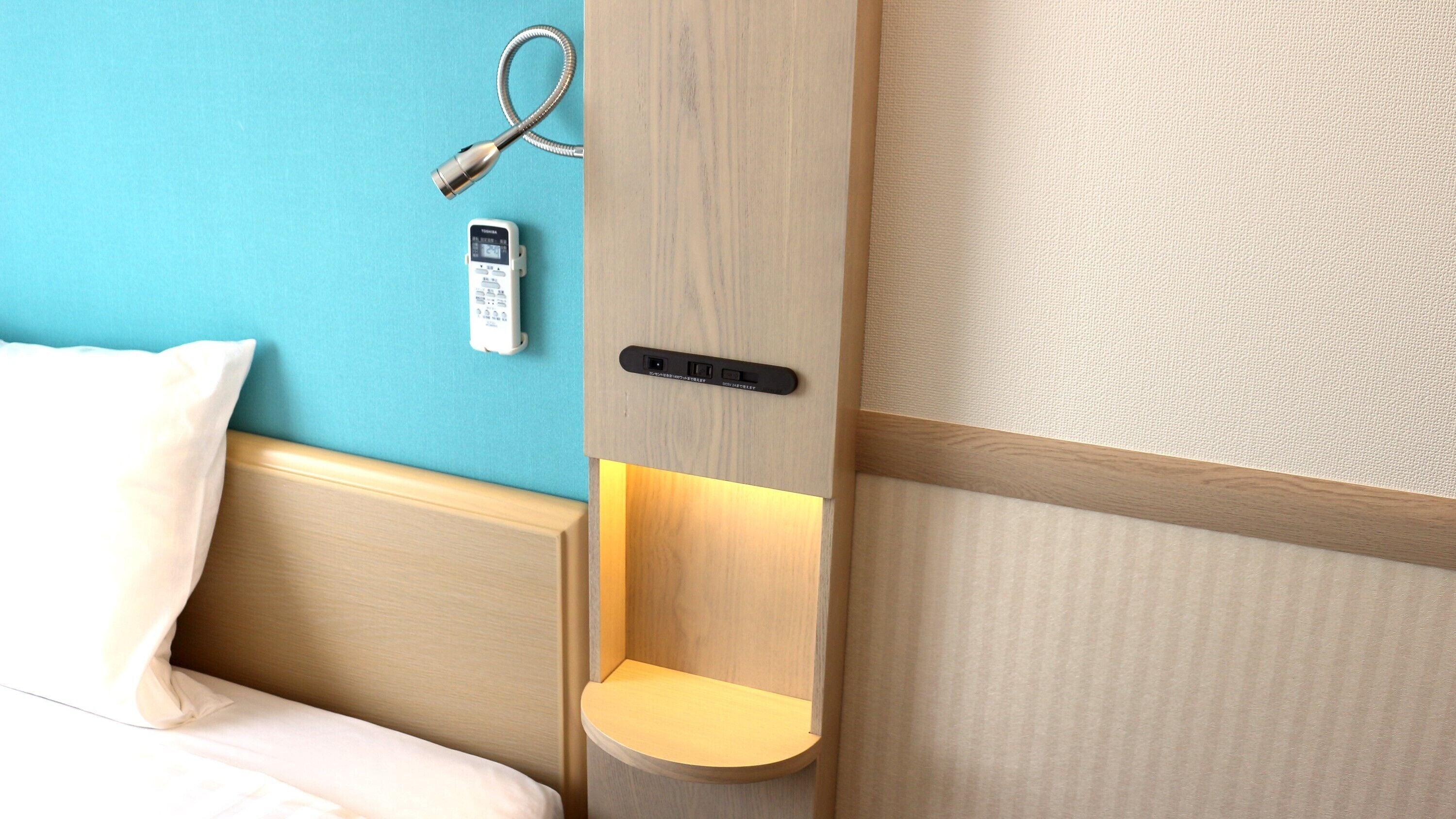 Une chambre d’hôtel avec une tête de lit en bois, une table de chevet avec une lampe, un téléphone fixé au mur et un panneau de commande.