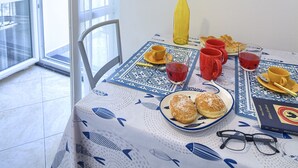 Une table à manger recouverte d’une nappe à motifs bleus et blancs, une assiette de pâtisseries, des tasses rouges et jaunes, un verre contenant une boisson rouge, une bouteille jaune et un livre.