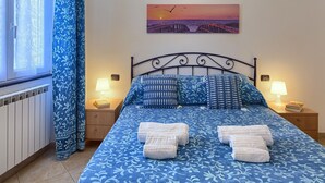 Un lit bien fait, avec une literie à motifs bleus et blancs, deux lampes de chevet et, au mur, un tableau représentant un coucher de soleil sur un pont.
