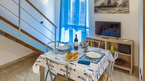 A dining area with a table set for two, a bottle of wine, and a bowl of fruit. A television is mounted on the wall above the table.