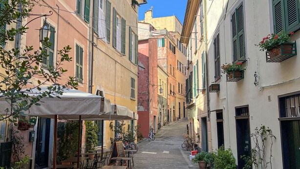 A narrow street with outdoor seating, buildings with green shutters, and potted plants.