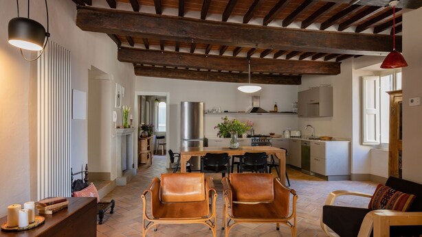 A spacious living area with a wooden ceiling, a dining table with chairs, a kitchen in the background, and a red pendant light.