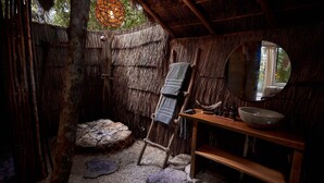 A rustic bathroom with a thatched roof, a wooden bench, a stone basin, and a mirror.