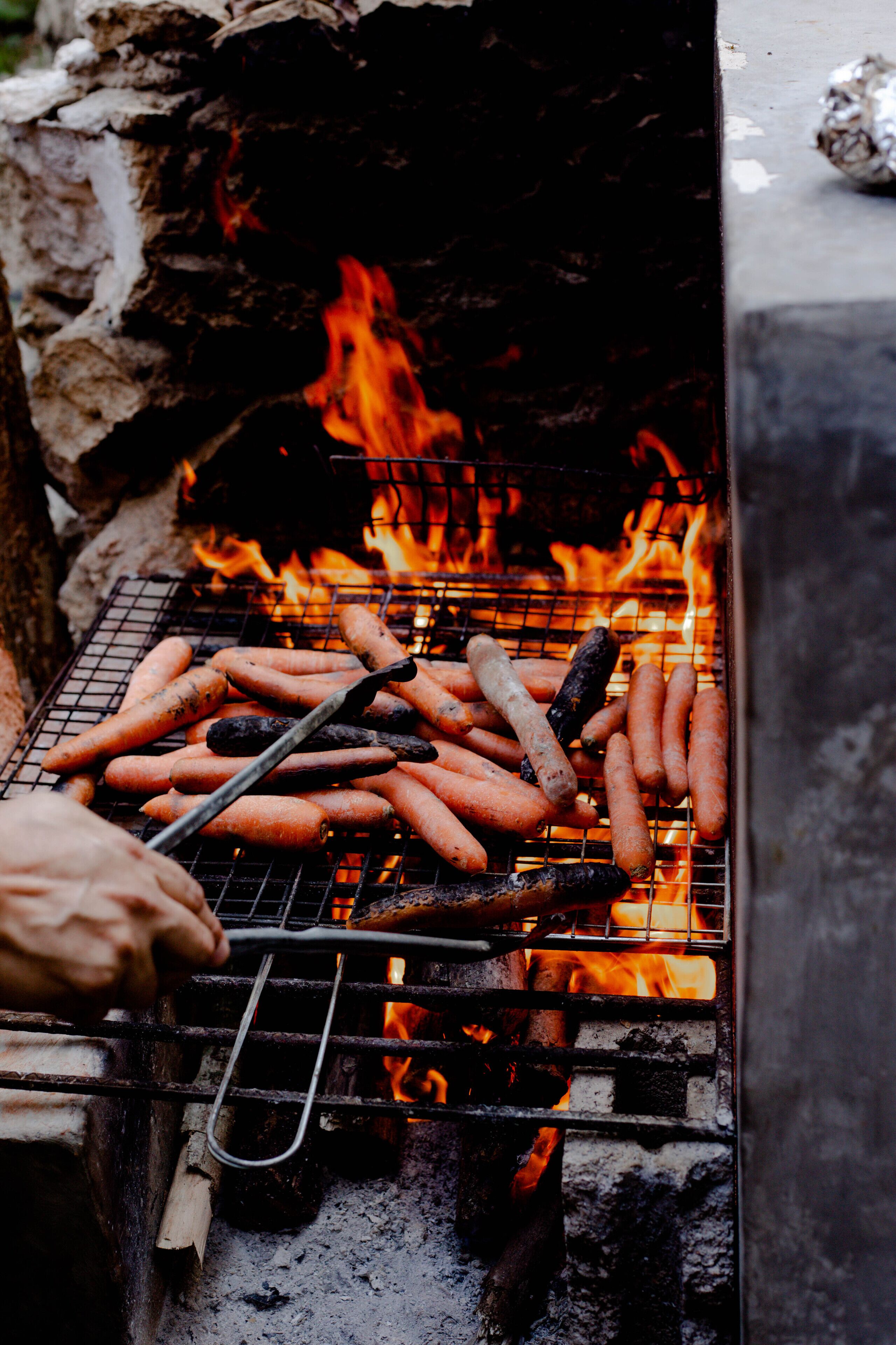 Una parrilla con salchichas y zanahorias asándose sobre fuego vivo.