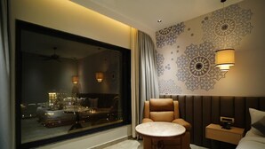 Una habitación de hotel moderna con un ventanal grande, un sofá, una mesa redonda, una cama y detalles decorativos en la pared.