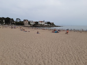 A sandy beach with people relaxing and sunbathing. There are houses in the background near the water.