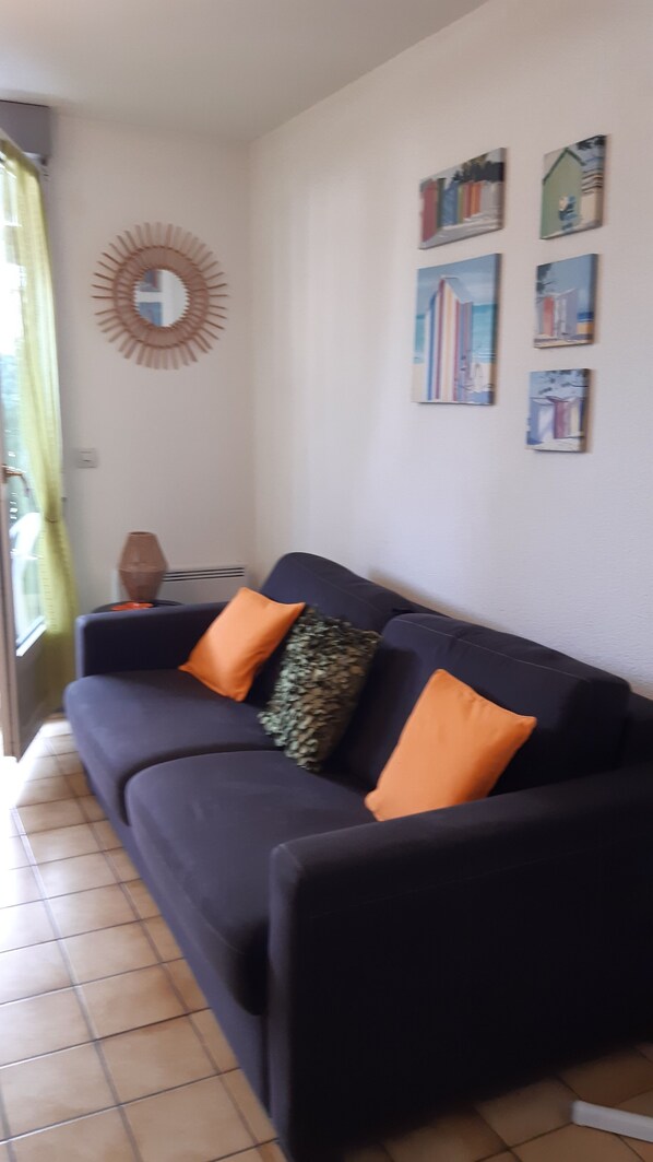 A dark-colored sofa with orange and green cushions, a round mirror, and several framed artworks on the wall.