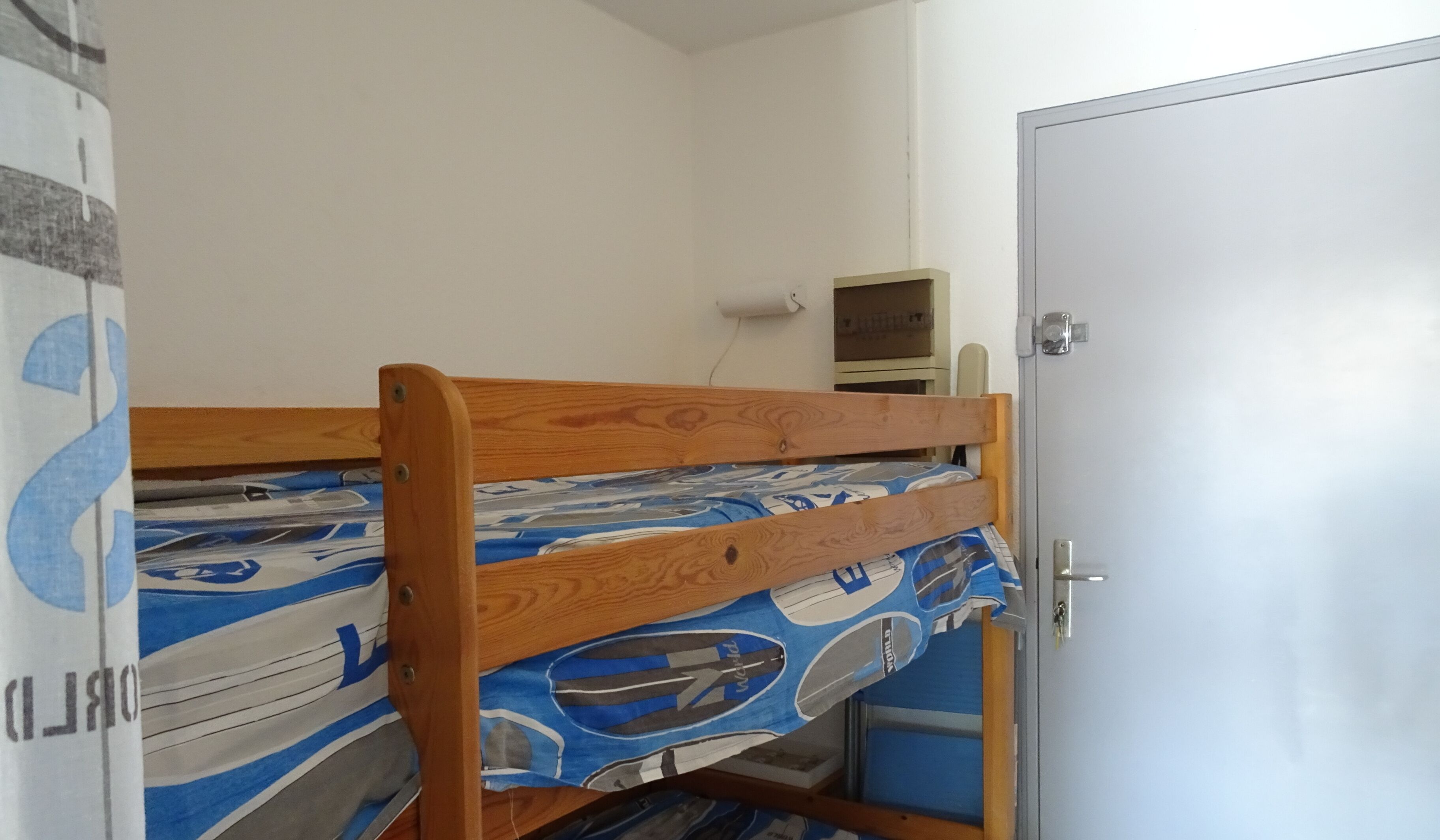 A bunk bed with a patterned bedspread, a desk with a computer, and a door in a room with white walls.