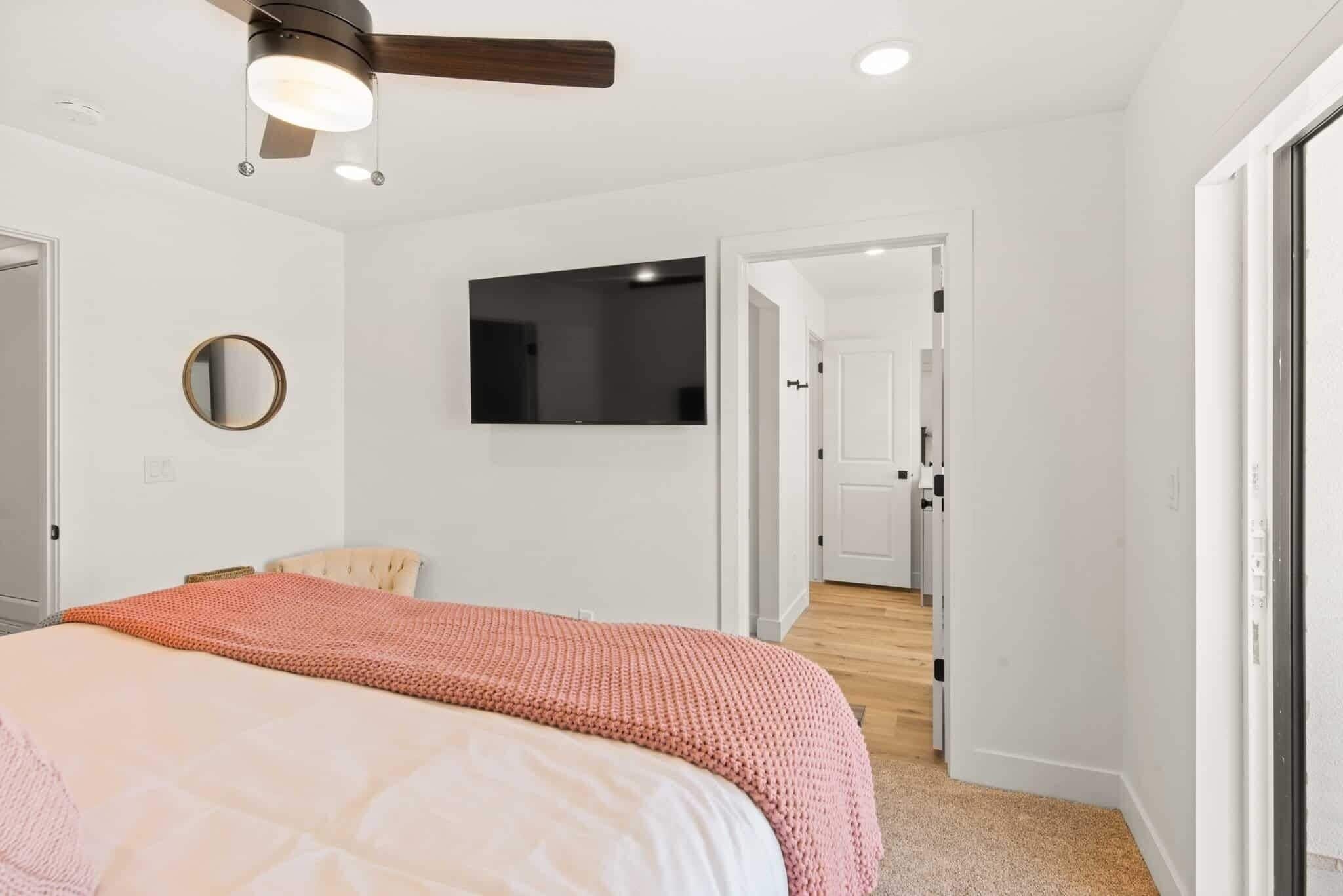 A bedroom with a bed, a ceiling fan, a TV mounted on the wall, and a door leading to another room.