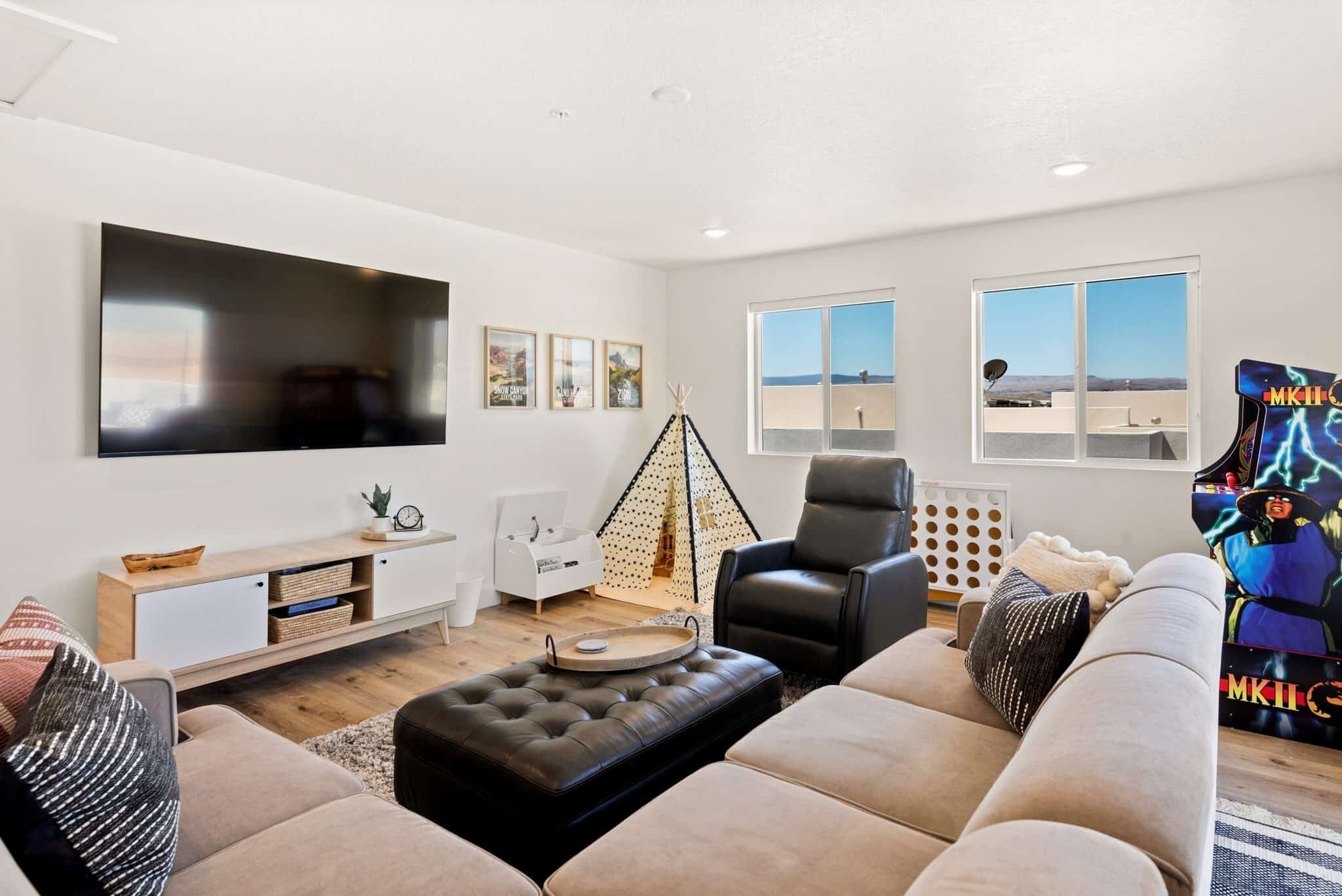 A modern living room with a large flat-screen TV, a sofa, a coffee table, and a gaming chair. There are framed pictures on the wall and a window with a view.