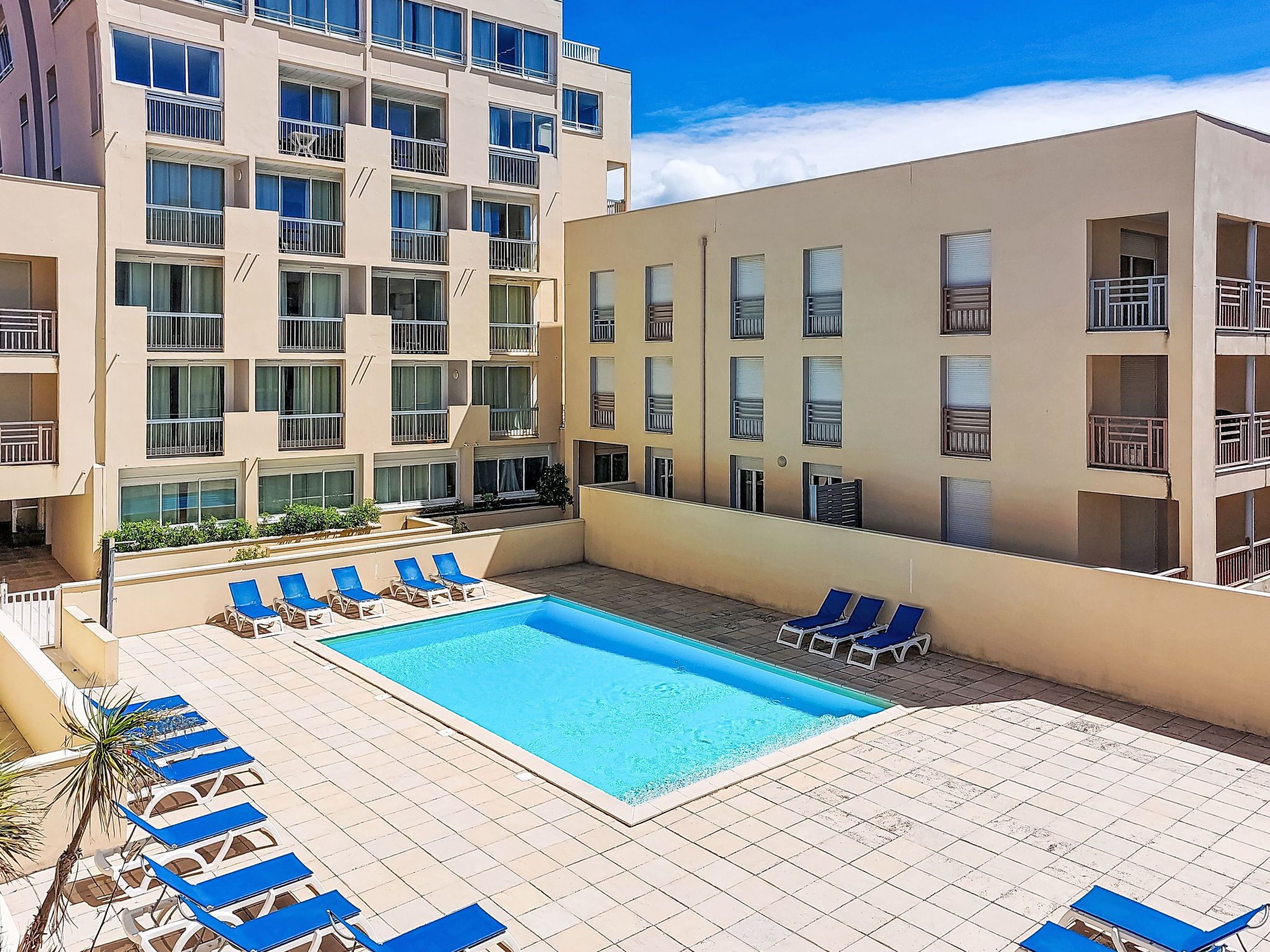 Un hôtel doté d’une piscine entourée de chaises longues, d’un immeuble à plusieurs étages avec des balcons, et d’un ciel d’un bleu limpide.