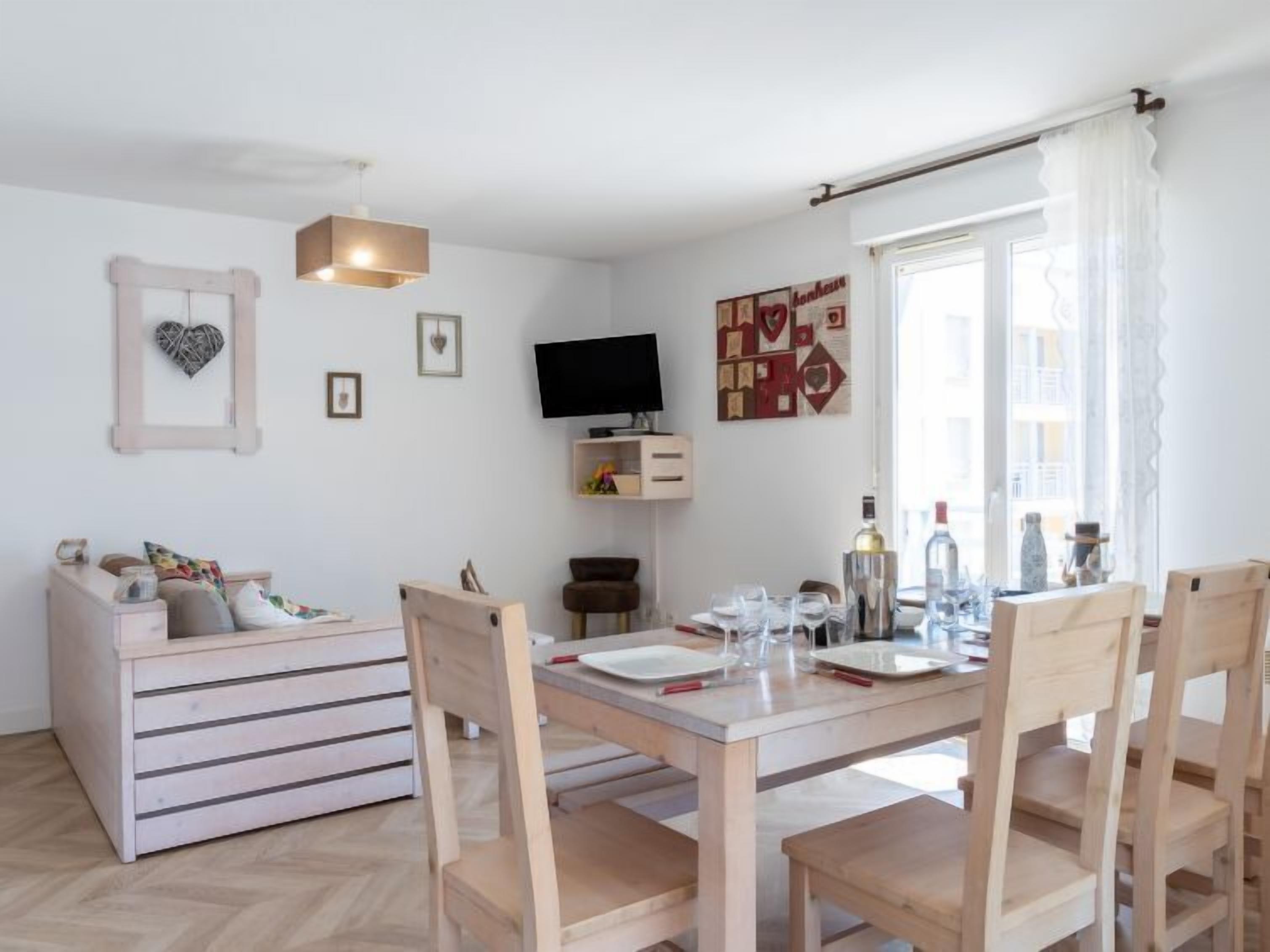 A modern living room with a wooden dining table, chairs, a sofa, and a TV on the wall.