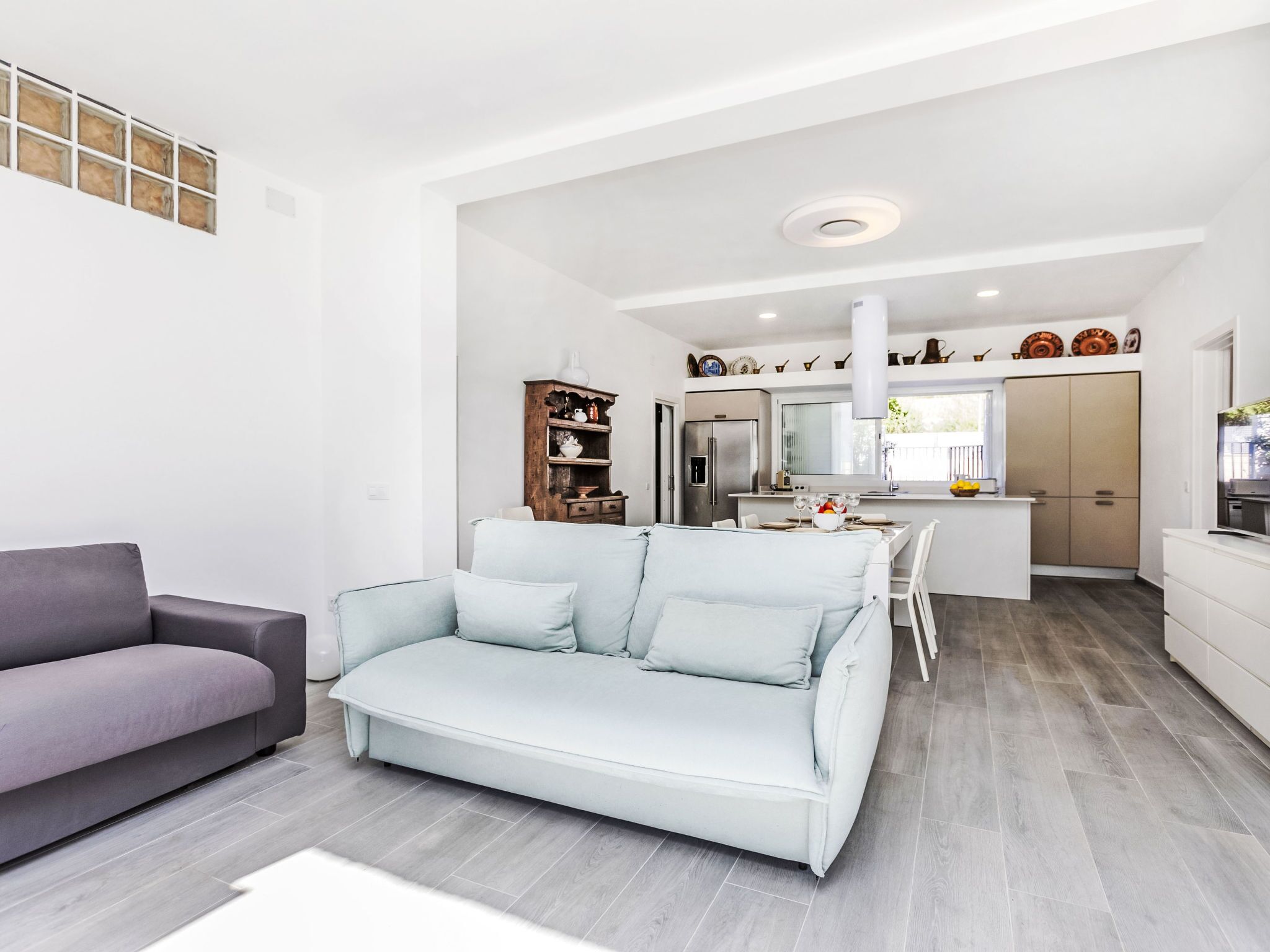 A modern living room with a grey sofa and a light blue sofa, a kitchen with white cabinets and a built-in oven, and a dining area with a white table and chairs.