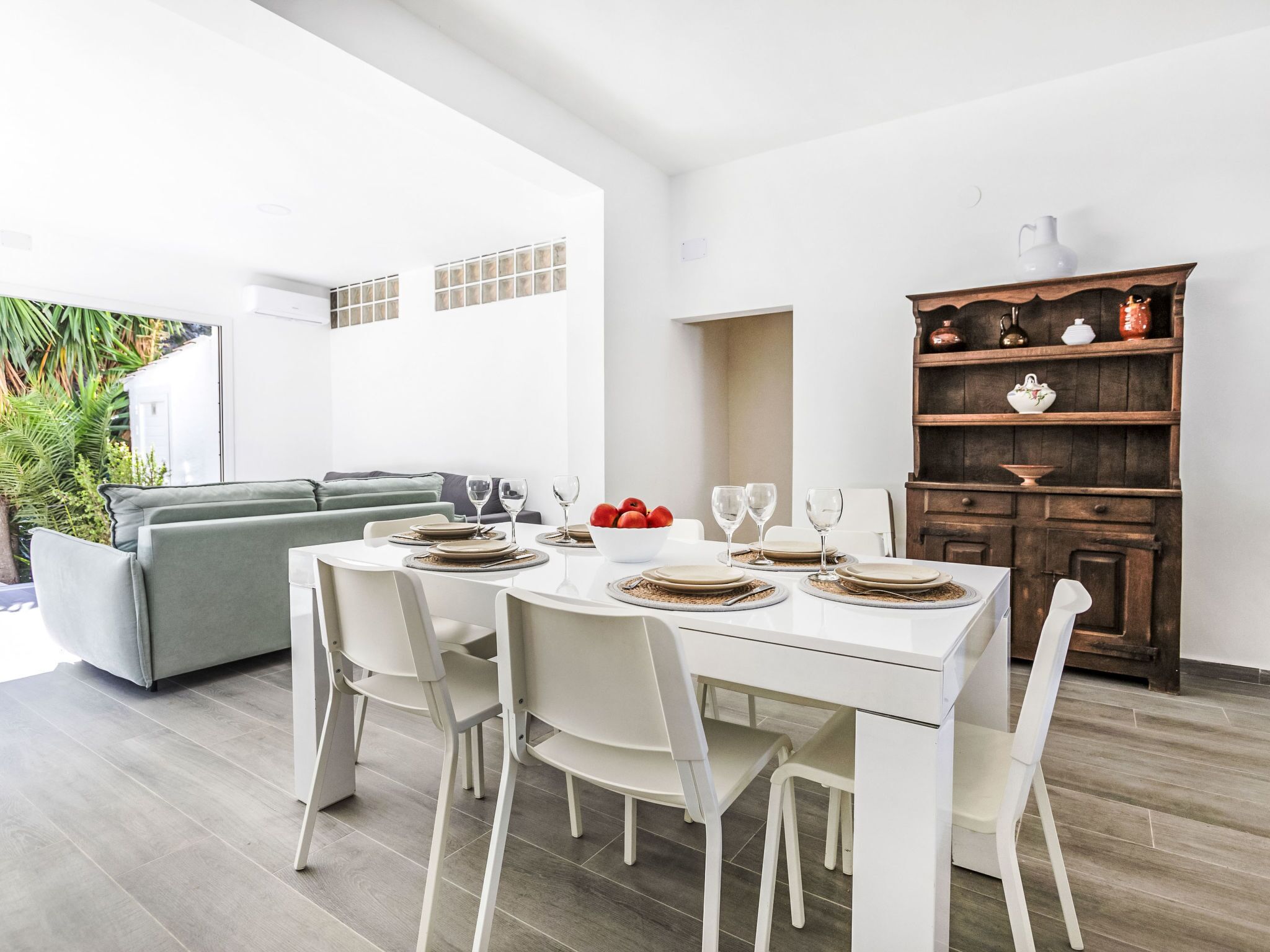 A dining area with a white table set for four, surrounded by white chairs. A gray sofa is visible in the background, and a wooden cabinet with decorative items is to the right.