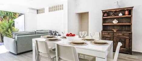 A dining area with a white table set for four, surrounded by white chairs. A gray sofa is visible in the background, and a wooden cabinet with decorative items is to the right.