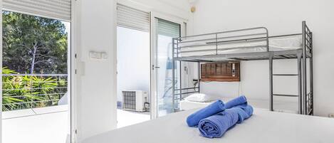 Habitación con literas, una colcha blanca, un par de toallas enrolladas y una ventana con persianas.