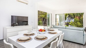 Un área de comedor moderna con una mesa blanca dispuesta para cuatro, un televisor de pantalla plana sobre una consola blanca, y vistas a un jardín exuberante a través de grandes ventanales.