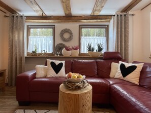 A cozy living room with a red leather sofa, two windows with curtains, a wooden beam ceiling, and a small wooden table with a basket. - Ehinger by Interhome (Eisenbach)
