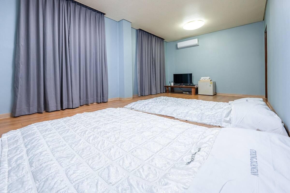 A hotel room with two beds, grey curtains, a flat-screen TV, a small table, and an air conditioner.