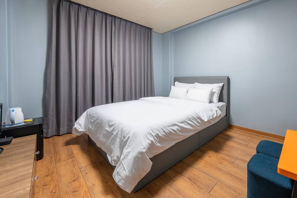 A hotel room with a bed, a nightstand, a coffee maker, a blue chair, and a small orange table.