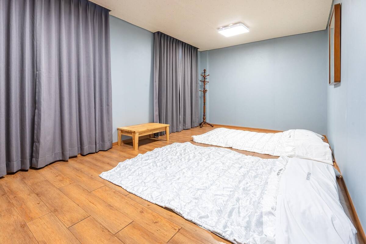 A room with wooden flooring, two beds with white bedding, grey curtains, a wooden bench, and a small table.