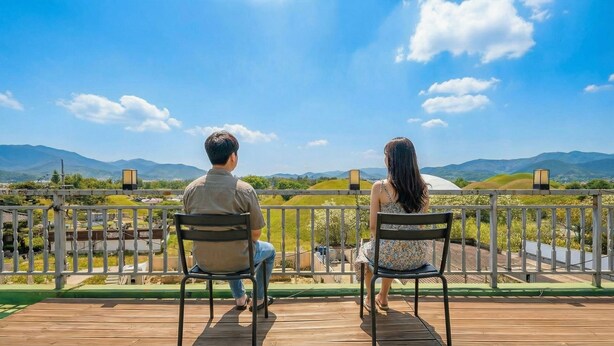 Two people seated on a wooden deck with a view of mountains and a clear sky. - Gyeongju Jang (Gyeongju)