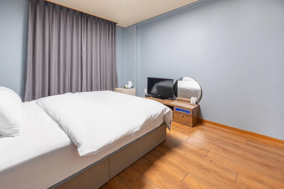 A hotel room with a bed, a TV, a bedside table with a mirror, and a wooden floor.