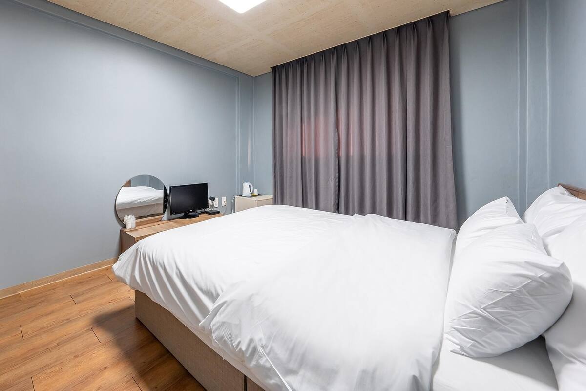 A modern hotel room with a large bed, a bedside table with a flat-screen TV, a mirror, and a window with curtains.