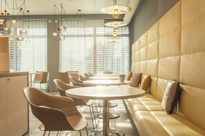 Un comedor moderno con mesas redondas, asientos acolchados, ventanas amplias con persianas e iluminación colgante.