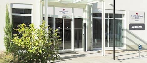 A hotel entrance with a glass door, a covered waiting area, and a 'Welcome' sign.