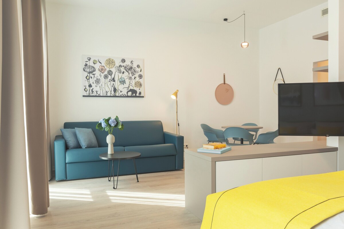 Una sala moderna con un sofá azul, una mesita redonda y una mesa de centro. Hay un cuadro en la pared y un televisor montado allí.