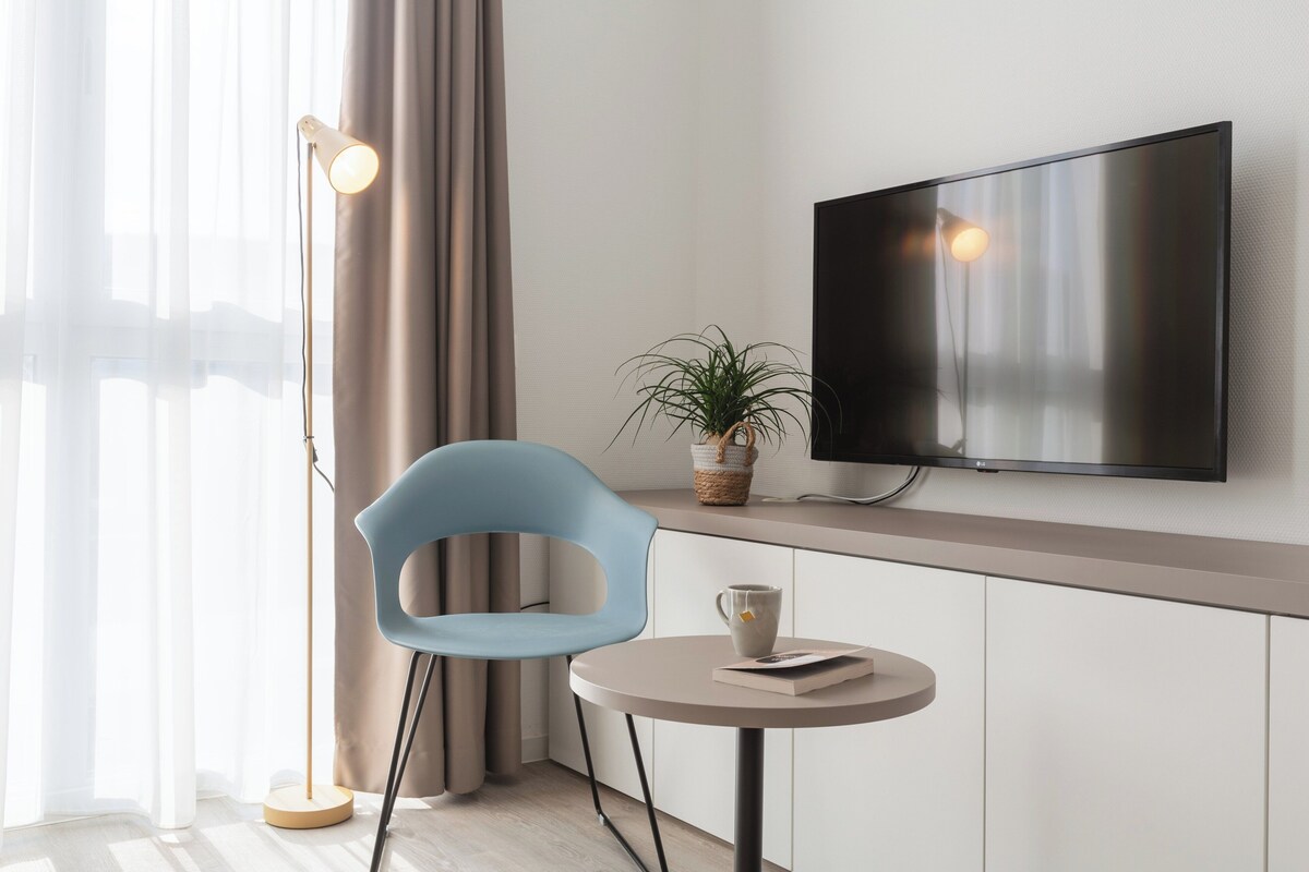 Una habitación de hotel moderna con una televisión de pantalla plana montada en la pared, una mesa redonda con un libro y una taza, una silla azul, una lámpara de pie y una planta en maceta.
