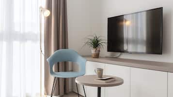 A modern hotel room with a flat-screen TV mounted on the wall, a round table with a book and a mug, a blue chair, a floor lamp, and a potted plant.