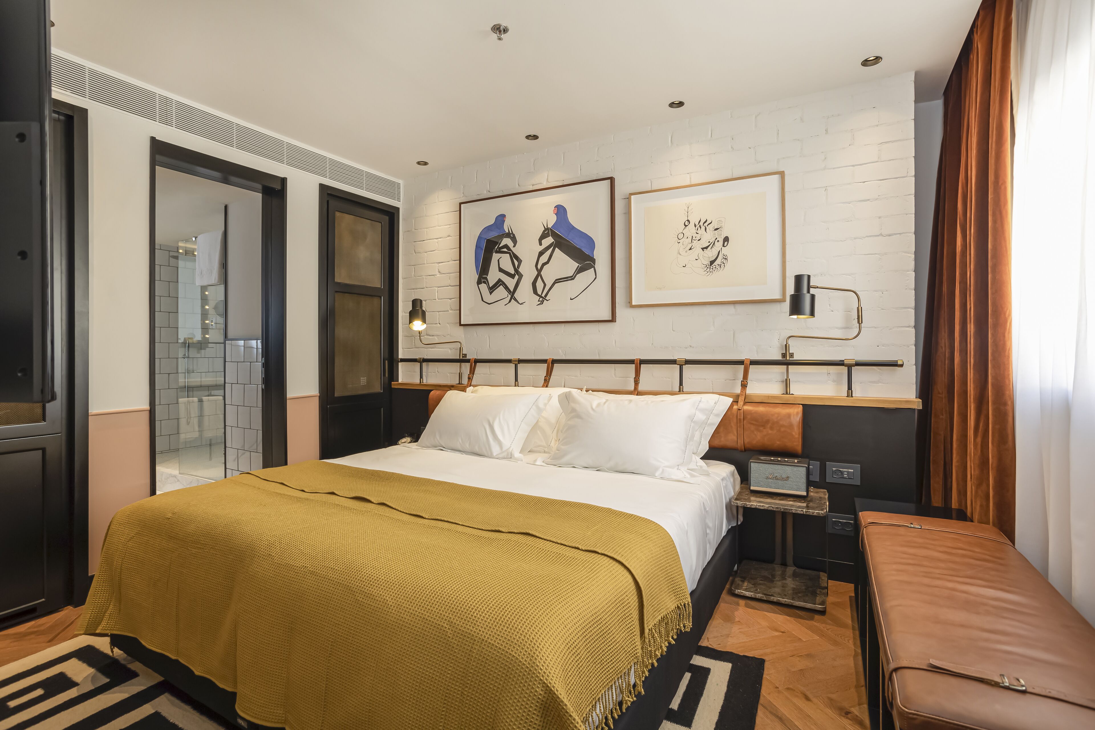 Une chambre d’hôtel moderne avec un grand lit, une table de chevet, un banc et des œuvres d’art accrochées au mur.