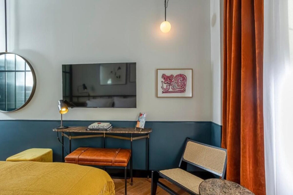 A room with a bed, a desk with a lamp, a chair, a television, and a framed artwork on the wall.