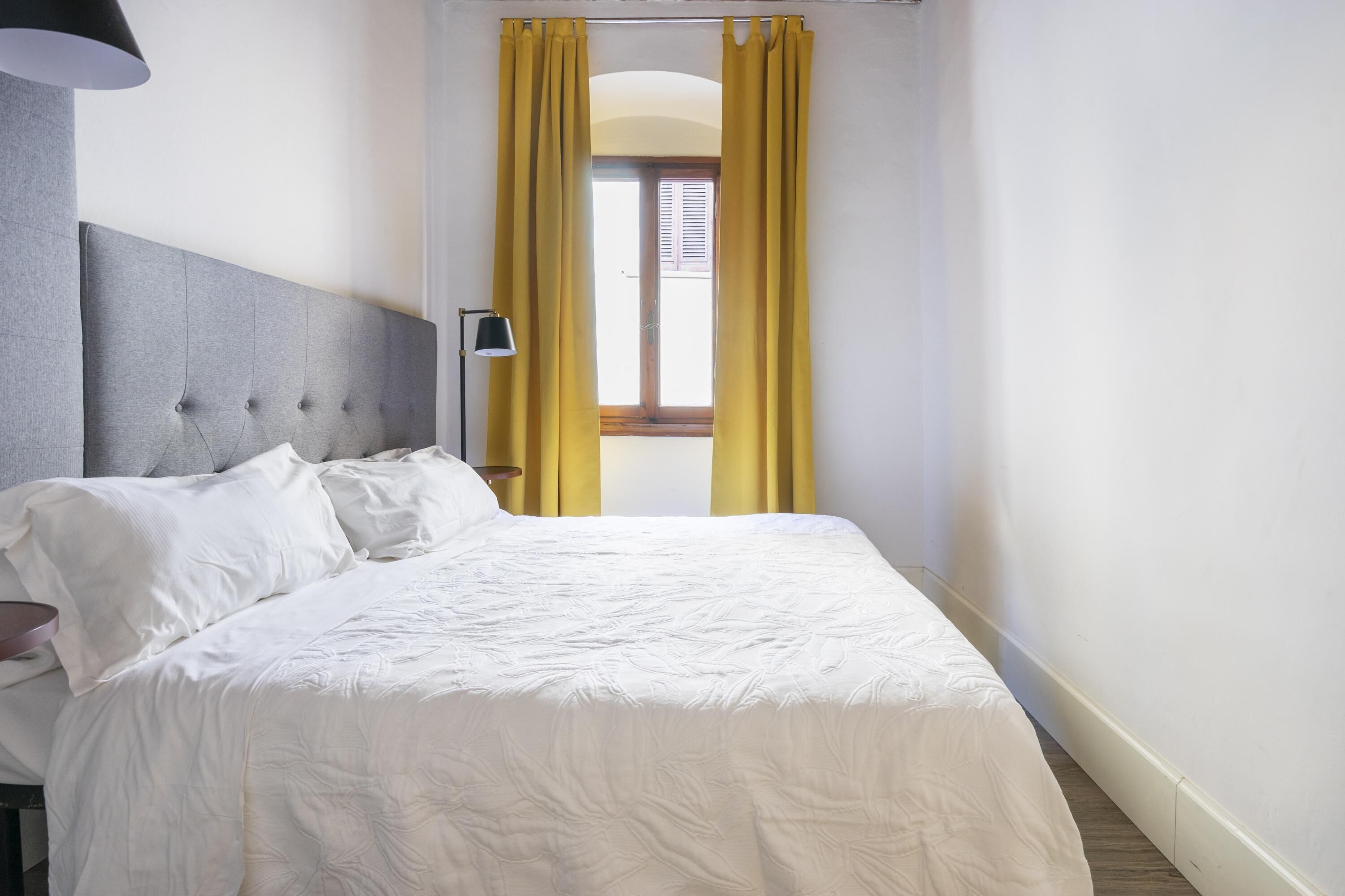 A neatly made bed with white bedding in a room with a grey headboard, a lamp on a bedside table, and yellow curtains on a window.
