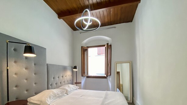 A bedroom with a tufted headboard, a bed with white linens, a bedside table, a floor lamp, a mirror, and a view of the outside through a window with wooden shutters. - Appartamento Ginori (Florence)