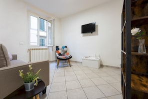 Quarto pequeno e bem iluminado com sofá, cadeira, televisão, radiador e planta em vaso.