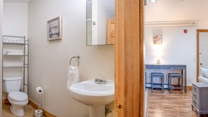 Een badkamer met toilet, wastafel en handdoekhouder. Een houten deur leidt naar een slaapkamer met een bed en nachtkastje.