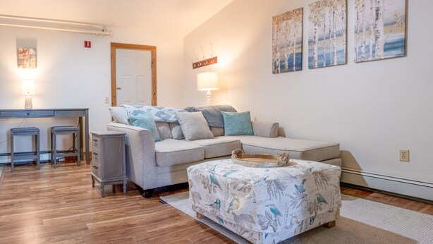 Een gezellige woonkamer met een bank, voetenbankje en twee bijzettafels. Er zijn wandlampen en een deur met een hendel.