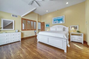 A bedroom with a large bed, wooden flooring, a ceiling fan, and a window with shutters.