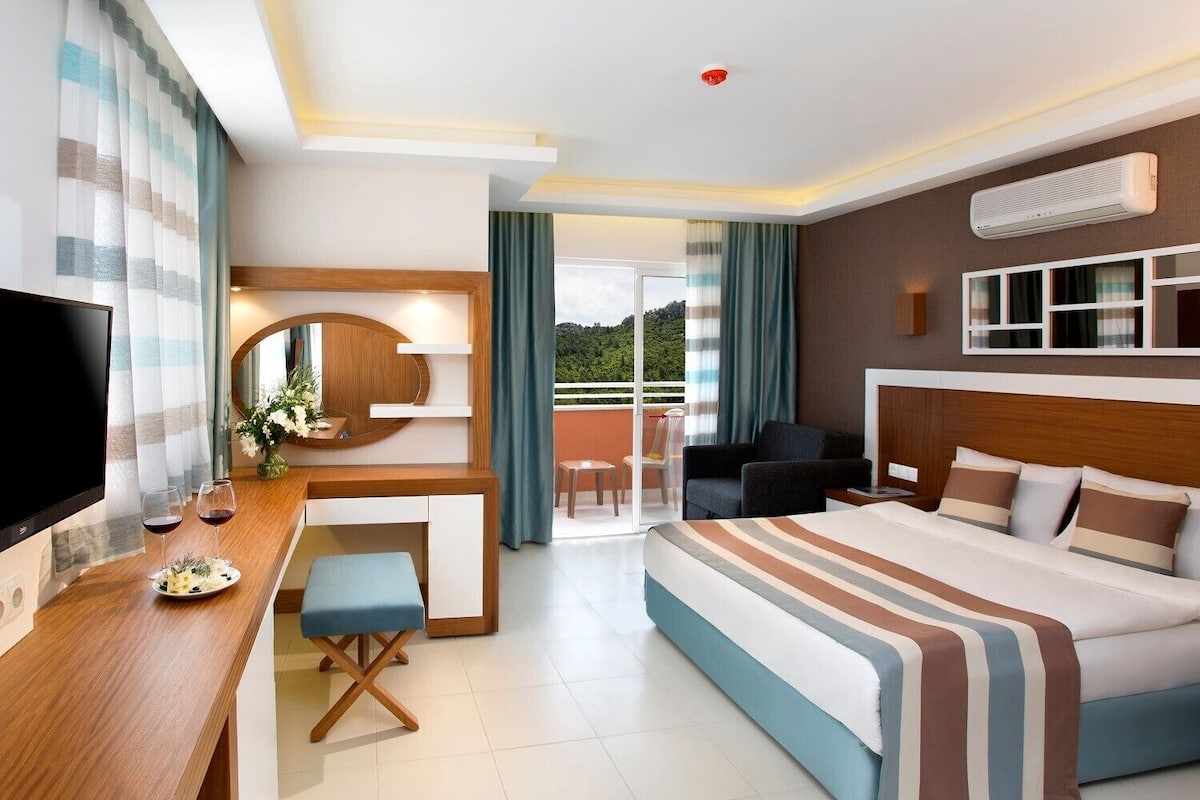 A modern hotel room with a large bed, a desk with a flat-screen TV, a small seating area, and a balcony with a view.