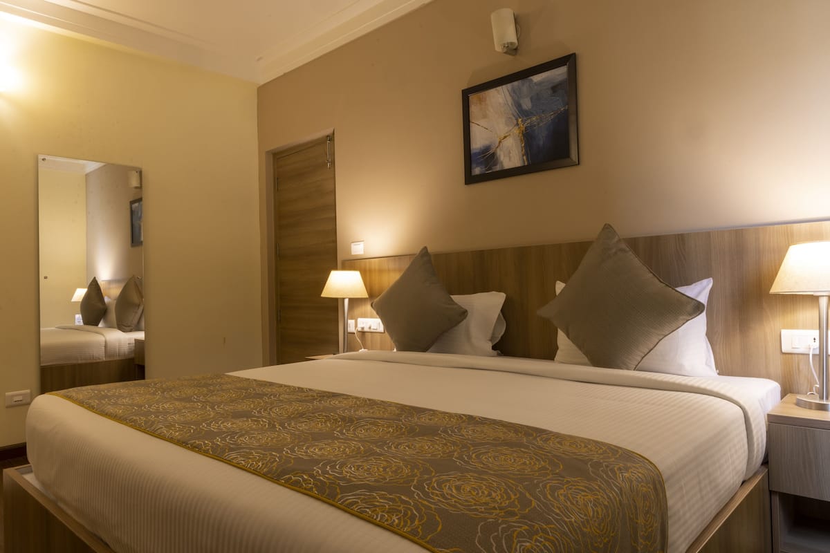 Quarto de hotel com uma cama grande, duas luminárias de cabeceira, um painel de madeira na cabeceira, um quadro na parede e um espelho.