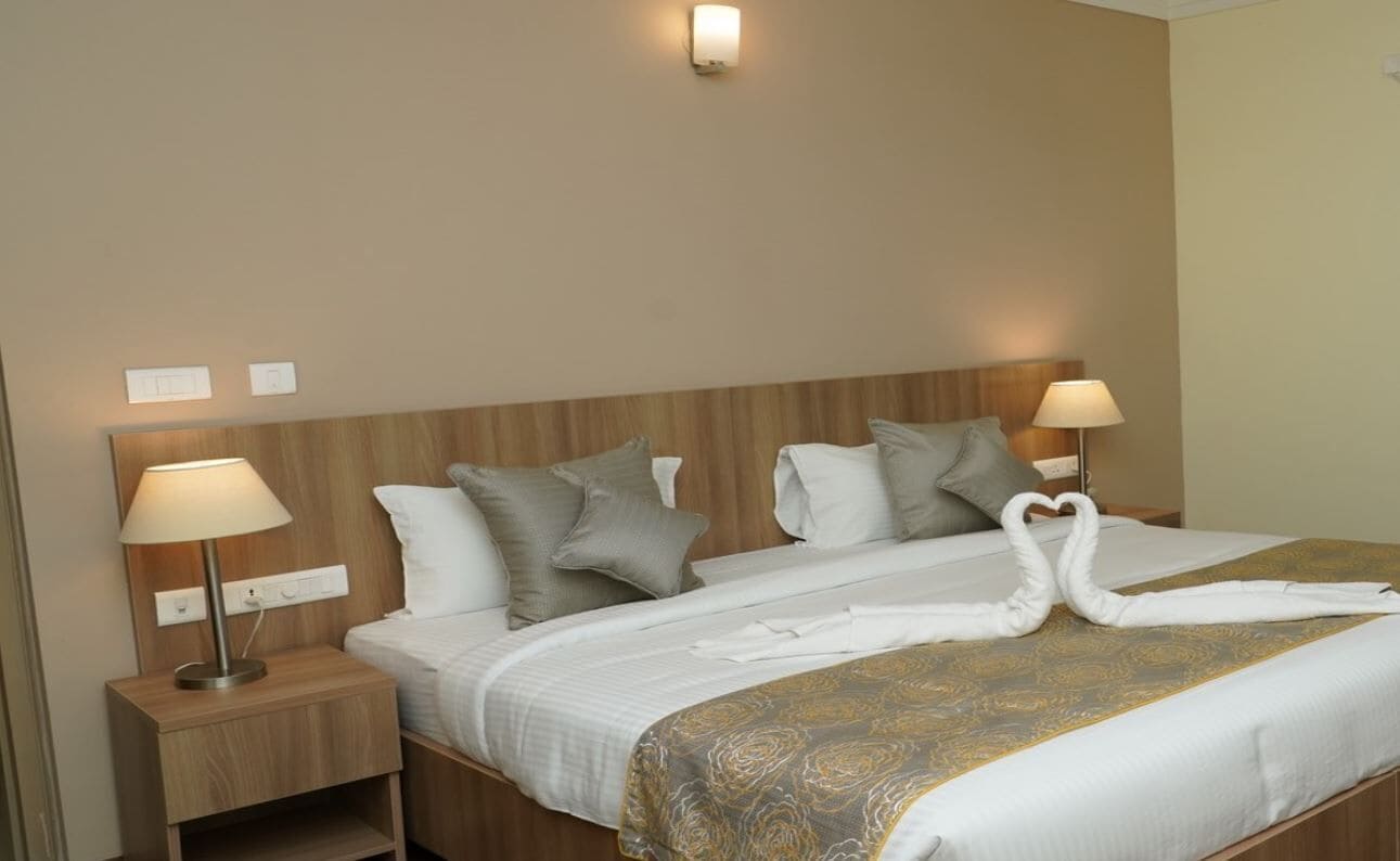 A hotel room with a wooden headboard, two bedside lamps, a bed with a white and gold patterned bedspread, and two towels arranged in a heart shape.